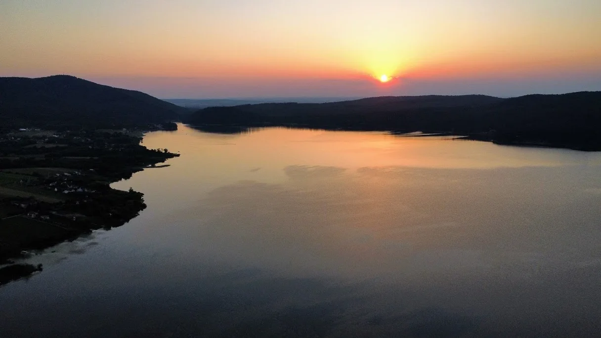 Apus peste Lacul Surduc, cu apa reflectând lumina portocalie între dealurile împădurite și malul cu sate din stânga.